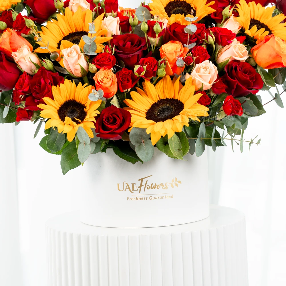A box arrangement of 6 sunflowers, 12 red roses, 3 red spray roses, 2 orange spray roses and 2 pink spray roses with green fillers.