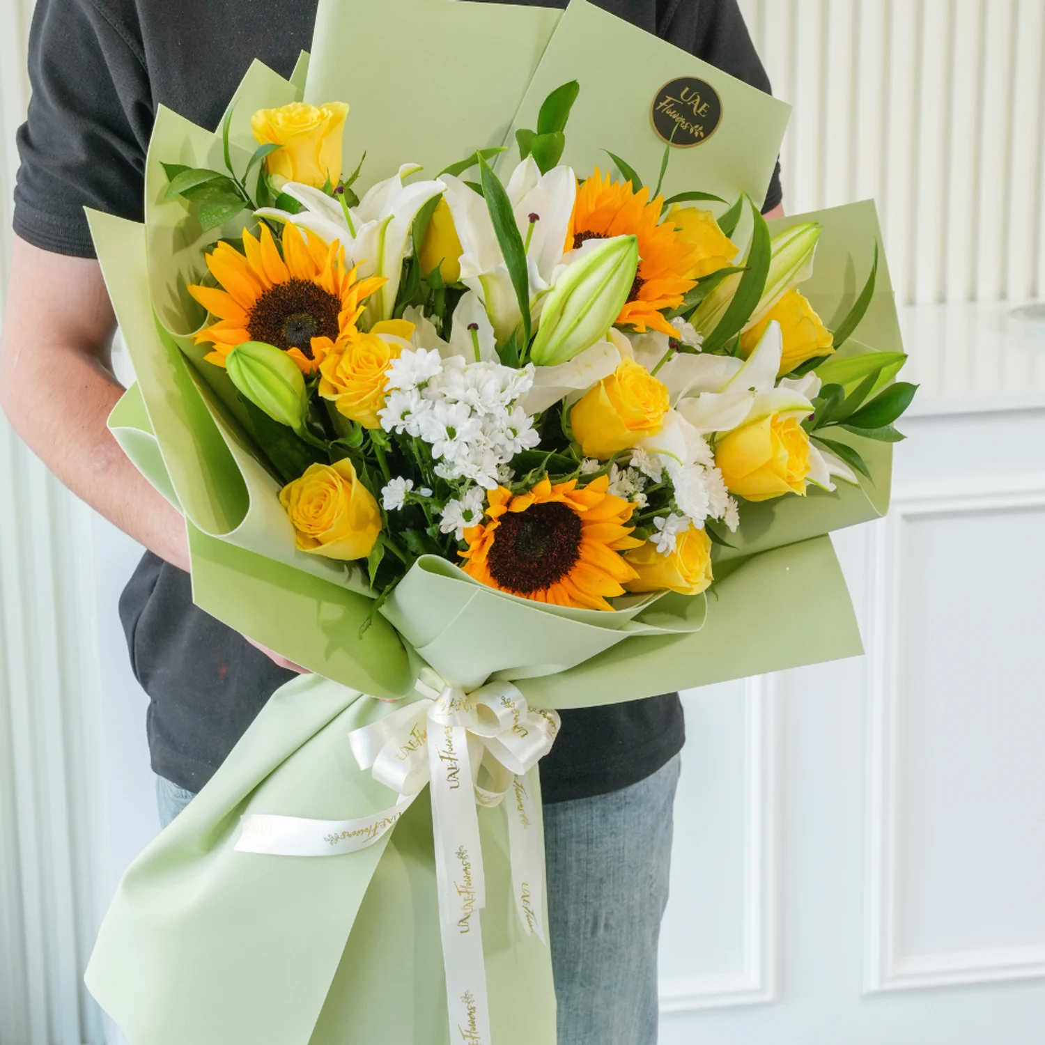 Bouquet of sunflowers, white lilies, yellow roses, and filler flowers wrapped in pastel green paper with a white satin ribbon – cheerful floral gift by UAE Flowers.