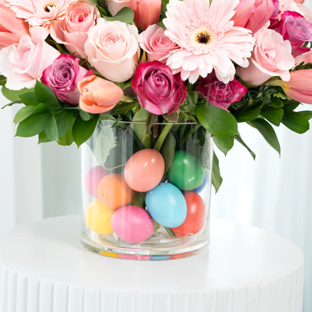 A vase arrangement of 3 pink gerberas, 7 purple roses, 15 pink roses, 10 pink tulips with green fillers and multi colored easter eggs.