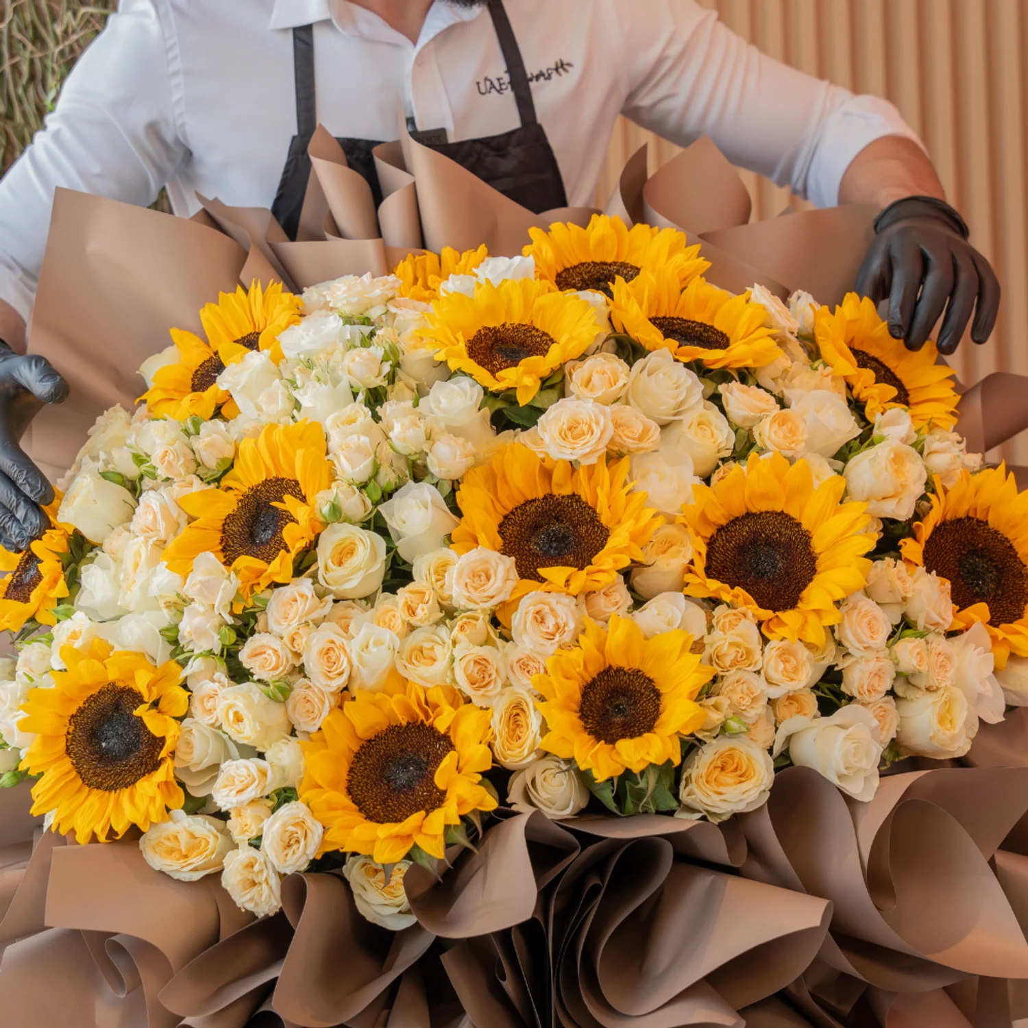 Tall luxury floral stand with 15 sunflowers, 50 spray roses, and 50 premium roses, wrapped in brown draping with satin ribbons – elegant event display by UAE Flowers.